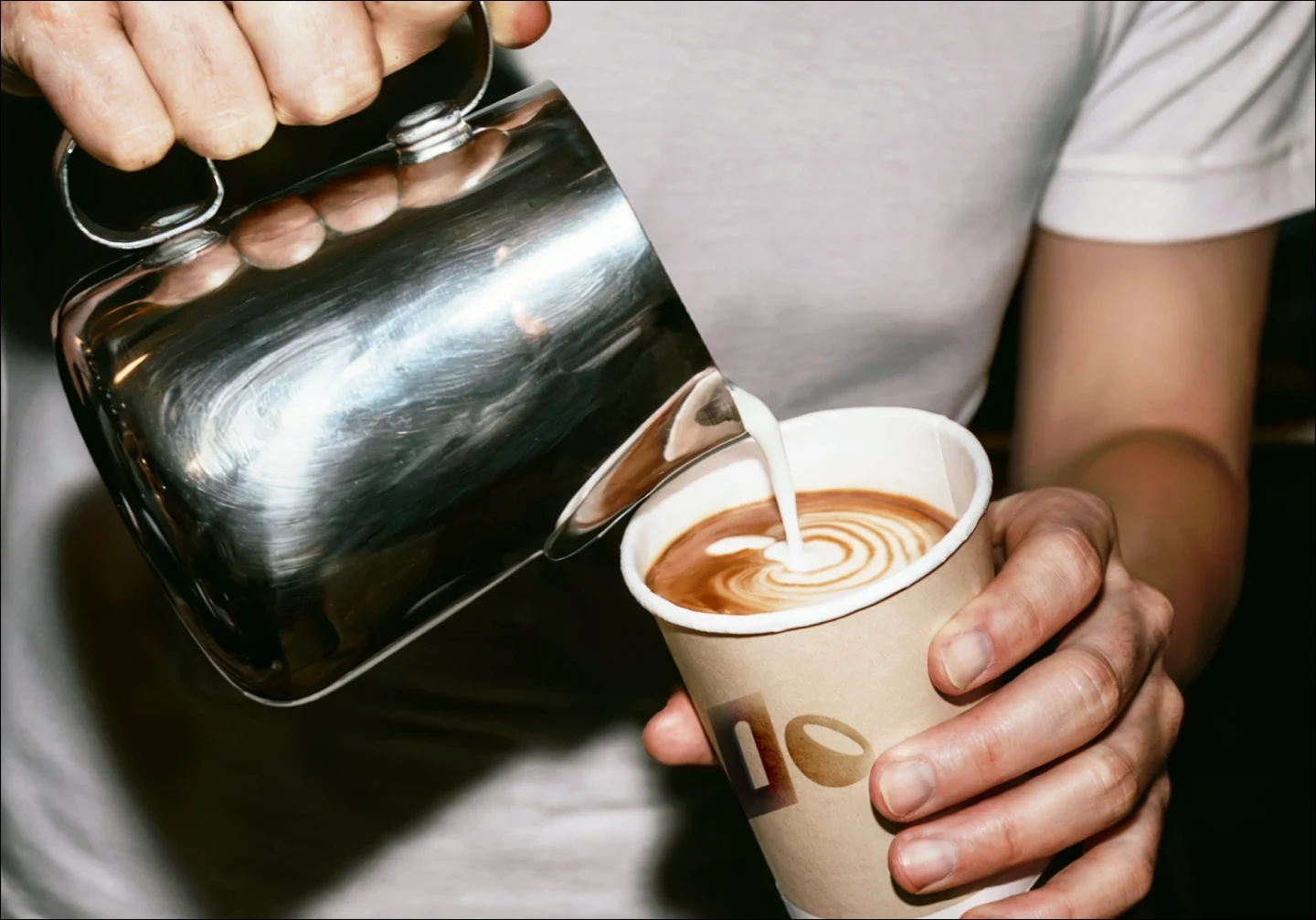 Barista pouring steamed milk into latte art in a Clay & Bean paper cup with logo branding visible.