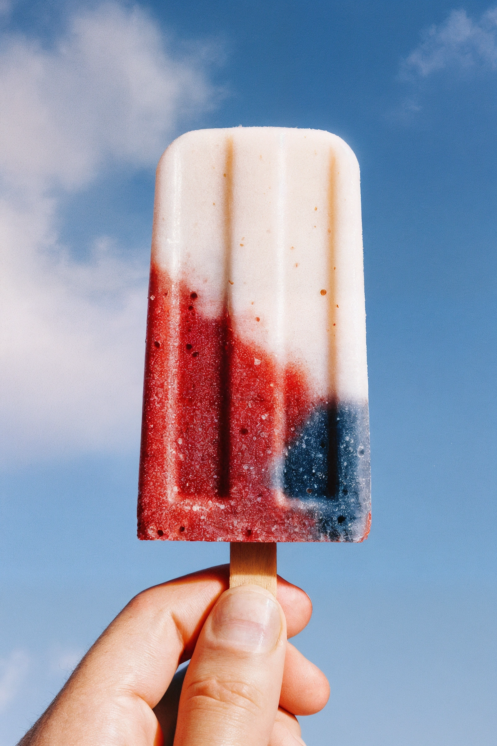A vibrant red, white, and blue popsicle held against a bright blue sky, evoking a refreshing summer vibe.


