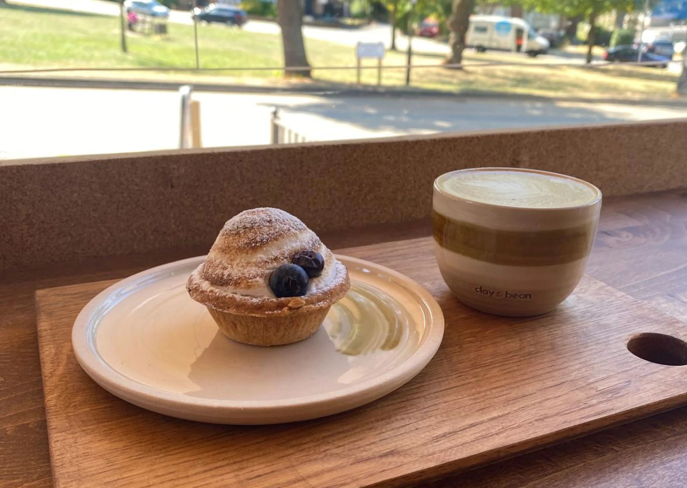Clay & Bean ceramic mug with latte served beside a powdered tart topped with blueberries on a wooden tray