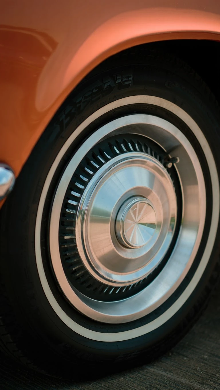 Close-up of a vintage car's chrome hubcap and whitewall tire, with an orange body reflecting light, showcasing classic automotive design.