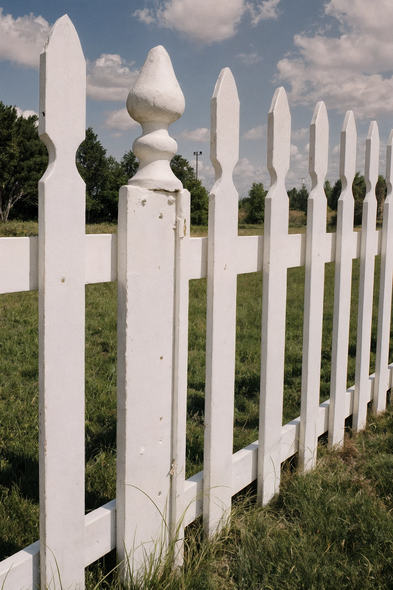 Close-up of a white picket fence with a decorative post cap, set against a grassy field and a partly cloudy sky.

