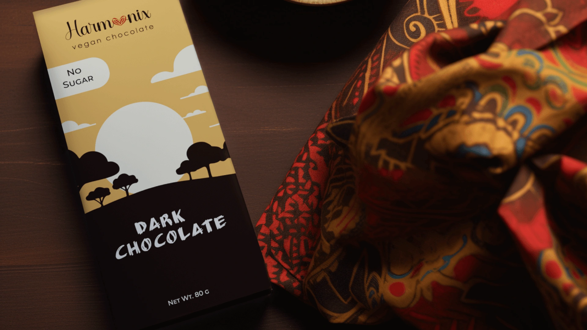The packaging design for the Harmonix Vegan Dark Chocolate is visually striking and appears to be crafted with the intent of celebrating the beauty of African nature and the traditional color palette of African cultures. The use of a warm and earthy color scheme creates an inviting atmosphere that aligns well with the natural and organic qualities associated with vegan products. The silhouette of the African landscape against a large sun suggests the origin of the cocoa and the traditional patterns imply a connection to the rich cultural heritage.