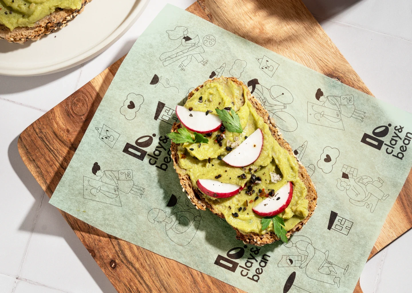 Avocado toast with radish and herbs on branded Clay & Bean wrapping paper with playful line art.